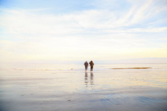 Couple standing on wet sand on beach and looking away. Man and woman relaxing on the sea ocean on sunset. Back view. Peaceful scene, calming waves, pastel cloudy sky, east coast. Happy time together.