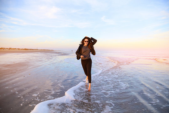 Smiling girl in hat running on beach on ocean coast. Female having fun in sea waves. Happy woman with long hair in leather jacket walking on wet sand and splashing water on sunset. Windy weather.