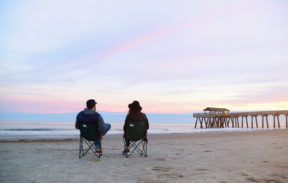 Couple sitting in deck chairs at beautiful sunset beach. Man and woman in hats and casual clothes relaxing near ocean pier jetty. Peaceful scene, calming waves, pastel cloudy sky, coast, wet sand.