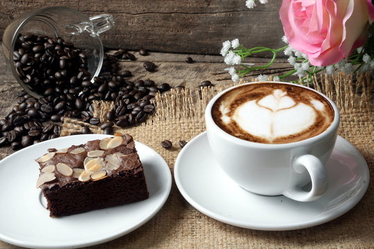 Cappuccino coffee and sweet chocolate brownies cake. A cup of latte, cappuccino or espresso coffee with milk put on a wood table with dark roasting coffee beans. Drawing the foam milk on top.
