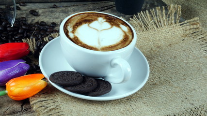 Cappuccino coffee and chocolate cookies. A cup of latte, cappuccino or espresso coffee with milk put on a wood table with dark roasting coffee beans and cookies. Drawing the foam milk on top.