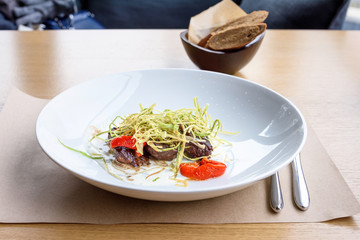 Beef medallions with greens, fry, red boiled pepper and white cheese sauce on a white plate, brown bread, restaurant table