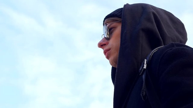 Male model in a black hooded coat black stylish glasses on the background of blue sky.