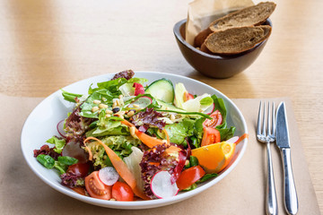 Close up photo of green healthy salad with ruccola, tomatoes, cucumbers, beet and seeds, brown bread, fork and knife on a wooden restaurant table