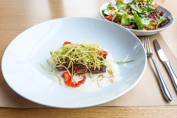 Beef medallions with greens, fry, red boiled pepper and white cheese sauce on a white plate, brown bread, restaurant table