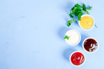 Classic set of sauces in white saucers: American yellow mustard, ketchup, barbecue sauce, mayonnaise. On cutting board light blue stone concrete table top view, copy space