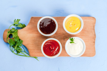 Classic set of sauces in white saucers: American yellow mustard, ketchup, barbecue sauce, mayonnaise. On cutting board light blue stone concrete table top view, copy space