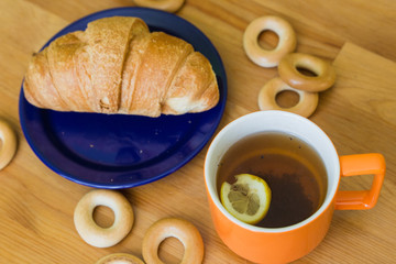 Mug full of black tea with lemon,  croissant on plate and small bagels on wooden table