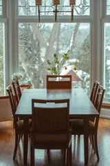 Large classic wooden dining table with  six chairs around standing by window, lemon tree and flowers on background, home interior decoration, toned with filters in retro vintage style.