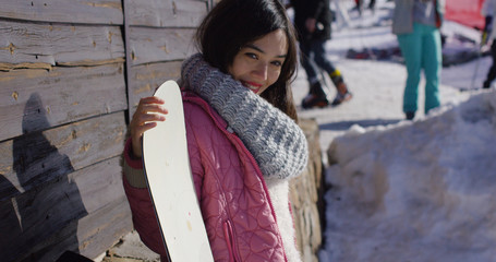 Smiling mixed race girl standing with snowboard in mountains resort. She looking to camra. Wearing woolen scarf and pink jacket.