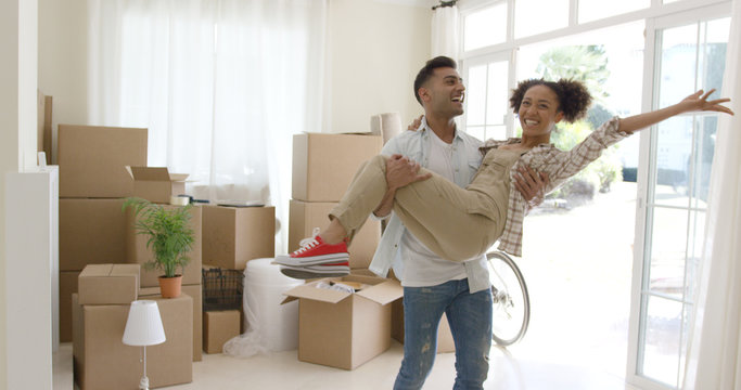 Ecstatic Young Couple Celebrating Their New Home With The Young Man Holding His Wife In His Arms As She Grins And Laughs At The Camera