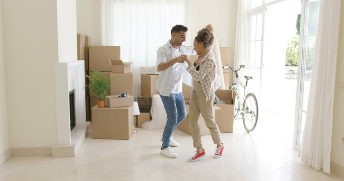 Happy young couple celebrating moving home dancing together in the living room surrounded by packing boxes