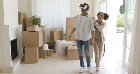 Cute young woman guiding her husband as he tries out a virtual reality headset in their new living room after moving house