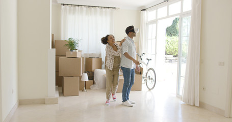Young man trying out virtual reality goggles guided by his wife standing in a bare living room surrounded by brown boxes during a move to a new home.