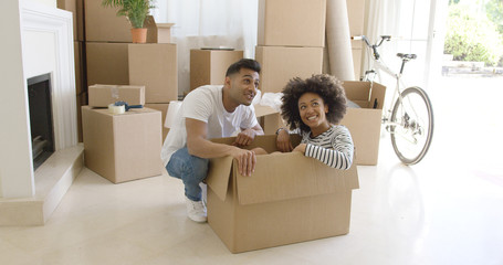 Happy young couple moving house together with the pretty young woman sitting inside a brown cardboard box with her husband crouching alongside smiling