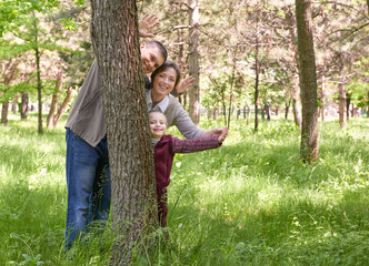 Fototapeta premium Happy family and child in summer park. People hiding and playing behind a tree. Beautiful landscape with trees and green grass