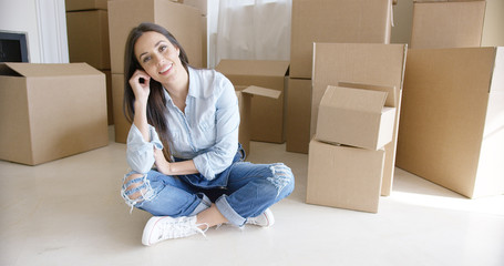 Happy young woman chatting on her mobile as she relaxes on the floor of the living room surrounded by boxes while moving home