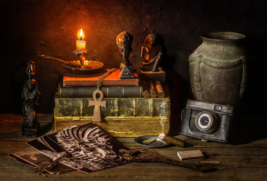 Classic still life with vintage books placed with old jar, illuminated candle, camera,old pictures,cigar and some ancient Egyptian sculptures on rustic wooden background.