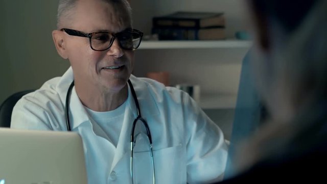 Positive professional middle-aged doctor talks about x-ray with patient at medical office. Medical man gives consultation about disease symptom and discuss prescription
