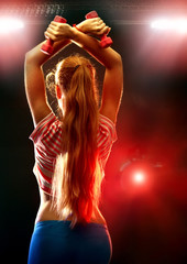 Naklejka premium Fitness girl exercising in gym with dumbbells. Woman has slim stomach works in multi-colored rays of sport room. Back view.