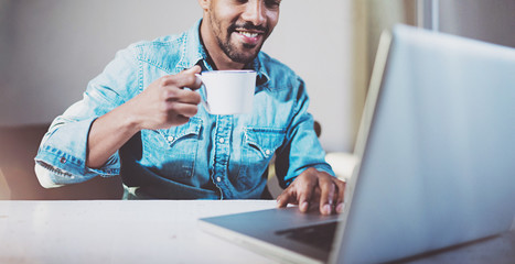 Smiling young African man making video conversation via modern laptop with friends while drinking black coffee in coworking office.Concept of happy business people.Blurred background,wide,crop.