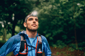 Fit male jogger with a headlamp rests during training for cross country trail race in nature park. - obrazy, fototapety, plakaty