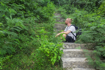 Naklejka na ściany i meble Man sitting on stairs in tropical forest. Side view of man sitting on a staircase in the jungle. Horizontal outdoors shot. 