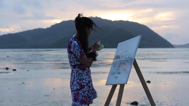 young girl draws a sunrise, on the beach at low tide. Work on the painting.