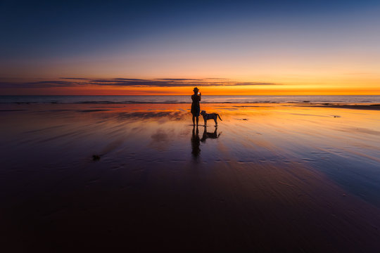 A woman taking a photograph of the sunset is silhouetted with her dog beside her. Western Australia, Australia.
