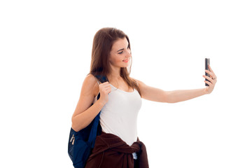 cheerful young brunette student girl with blue backpackand and mobile phone in her hands makes selfie isolated on white background