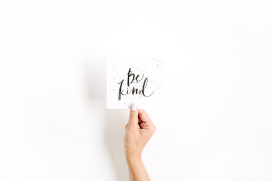 Minimal pale composition with girl's hand holding card with quote Be Kind written in calligraphic style on paper on white background. Flat lay, top view