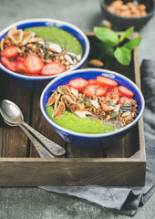 Healthy breakfast. Green smoothie bowls with strawberries, granola, chia and pumpkin seeds, dried fruit and nut in wooden tray over grey concrete background, selective focus. Vegetarian food concept