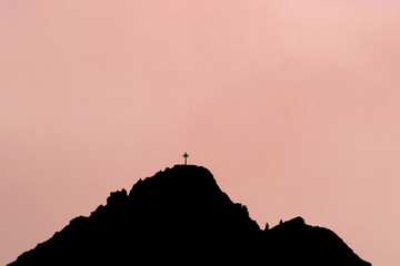 Black silhouette of mountain peak with summit cross. Rose background.