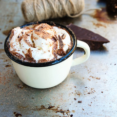 Vintage mug of hot chocolate cocoa with whipped cream and slice of bitter chocolate on vintage metal background. Winter times drink concept. Delicious cold weather beverage. Copy space.