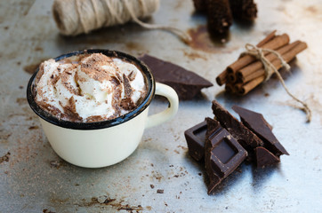 Enamel mug of hot chocolate cocoa with whipped cream, slice of bitter chocolate and cinnamon sticks on vintage metal background. Winter times drink concept. Delicious cold weather beverage.