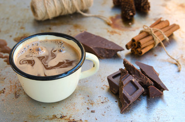 Enamel mug of hot chocolate cocoa, slice of bitter chocolate and cinnamon sticks on vintage metal background. Winter times drink concept. Delicious cold weather beverage.