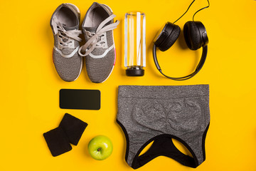 Fitness accessories on yellow background. Sneakers, bottle of water, headphones and sport top.