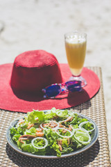 Seafood salad, orange fresh juice, hat and sunglasses on the table near sea