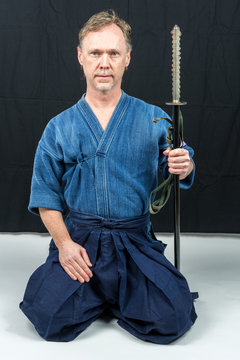 Caucasian male training Japanese sport, iaido. Sitting on floor holding a Japanese sword looking at camera. Indoor shot with black background.