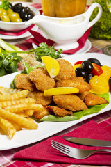 Breaded and deep fried tilapia bites on a white plate with French fries, lemon, polenta, cherry tomatoes and olives.