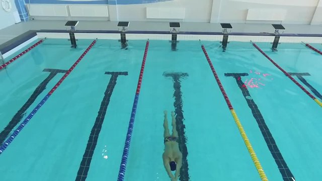 Flight over male swimmer swims in a pool HD aerial video. Freestyle training: turns underwater dolphin kick. Professional athlete on water lane