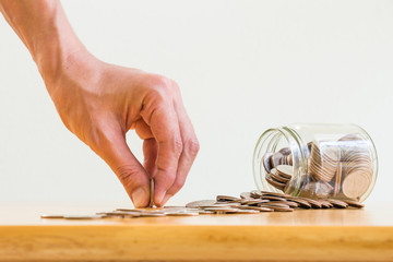 Business concept, saving planning with coins in glass jar and the man's hand holding one coin on wooden table over white wall background with space