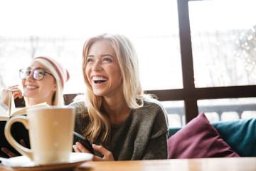 Laughing two women friends sitting in cafe and reading book