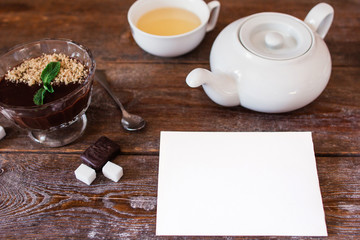 Blank card with teapot and dessert on table. Top view on wooden table with set for drinking tea and chocolate cream with white empty sheet of paper, free space