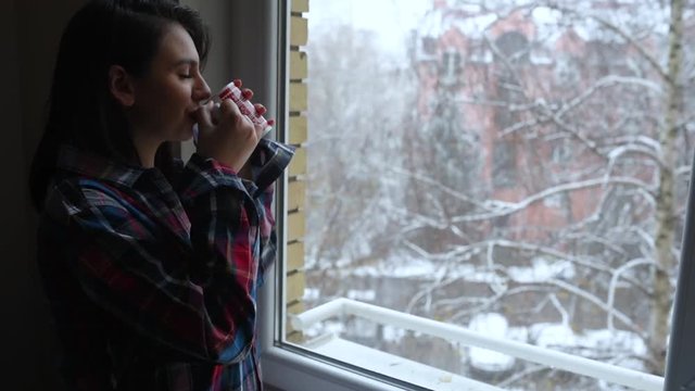 Young girl in plaid shirt drinking hot tea and watching snowfall