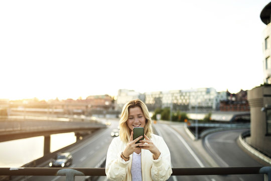 Happy young woman using mobile phone while standing on bridge against sky