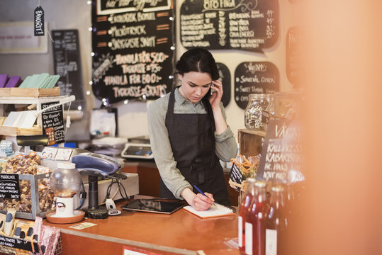 Owner talking on phone while writing on note pad at checkout counter in store - Powered by Adobe