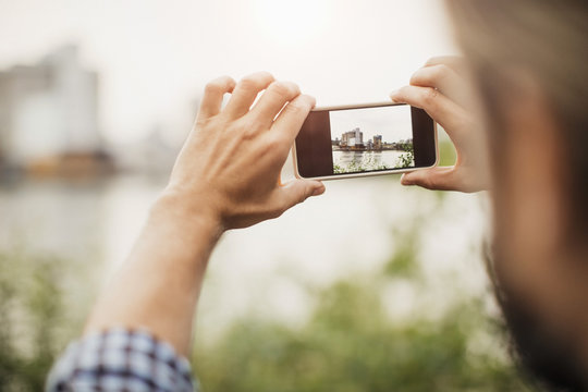 Cropped image of man photographing silo through smart phone against river