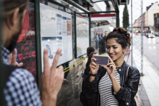 Smiling woman photographing man with smartphone at bus stop