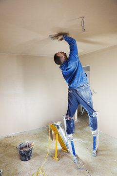 A plasterer wearing stilts smoothing fresh plaster high up on the walls of a house under construction. 